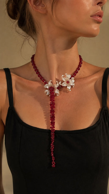 Elegant woman wearing a classic black top, styling a dramatic red beaded lariat necklace featuring a large sculpted white iris flower.